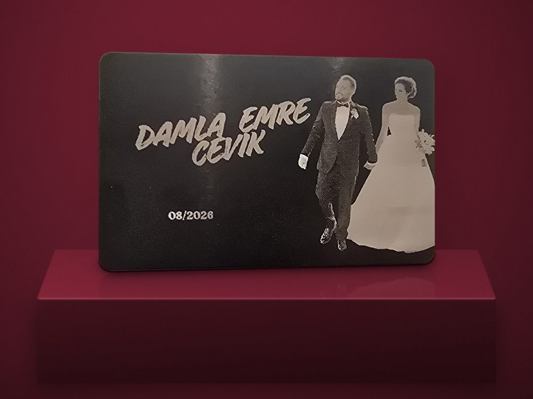 Black engraved metal wedding card for Damla & Emre Çevik with the date 08/2026, catching the light on a burgundy stand